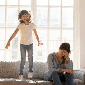 Niña hiperactiva brincando en el sofá mientras madre frustrada se cubre el rostro — imagen representativa del TDH en niños.